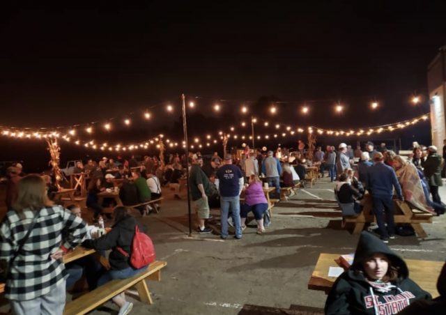 People gathered at picnic tables with lights strung above