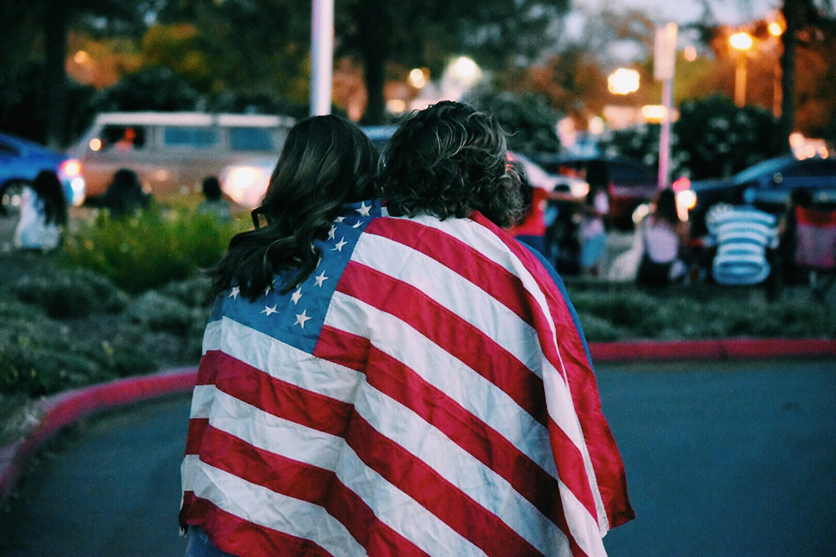 2 women wrapped in an United States flag watch at the edge of the celebrations on the 4th of July.