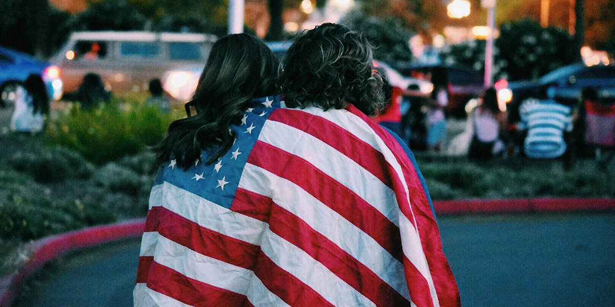 2 women wrapped in an United States flag watch at the edge of the celebrations on the 4th of July.