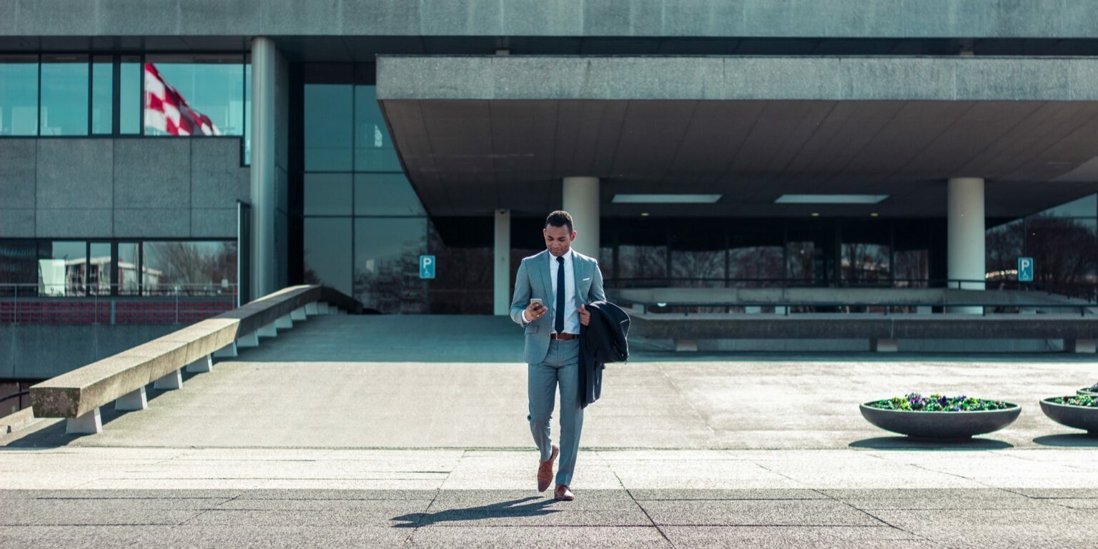 A man wearing a grey suit checks his phone while walking