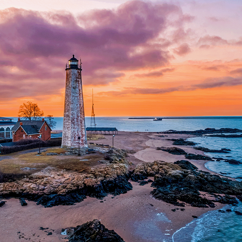 Sunrise behind a lighthouse on the rocky and sandy shores of Connecticut.